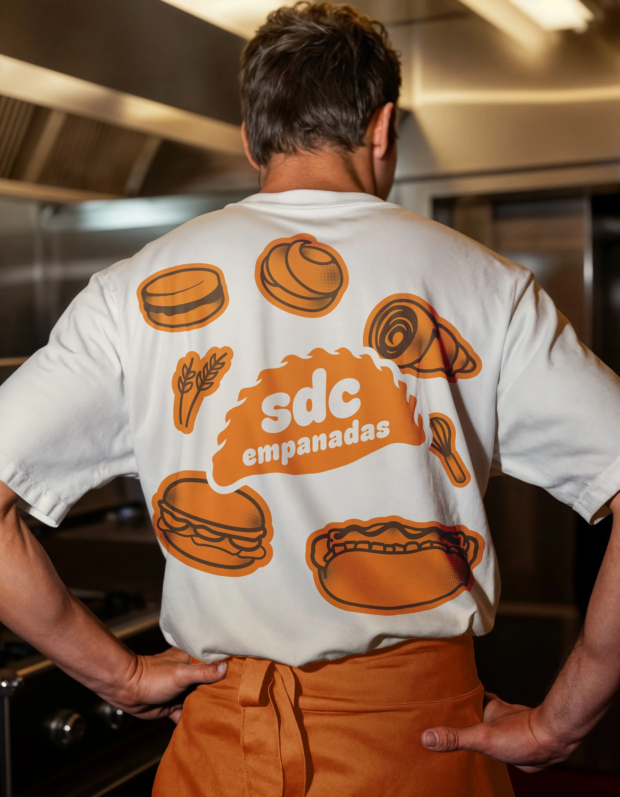 Person wearing a white t-shirt with orange food cartoon illustrations and text, standing in a kitchen. The illustrations include a burger, a chicken drumstick, and a slice of meat, along with wheat and a fork.