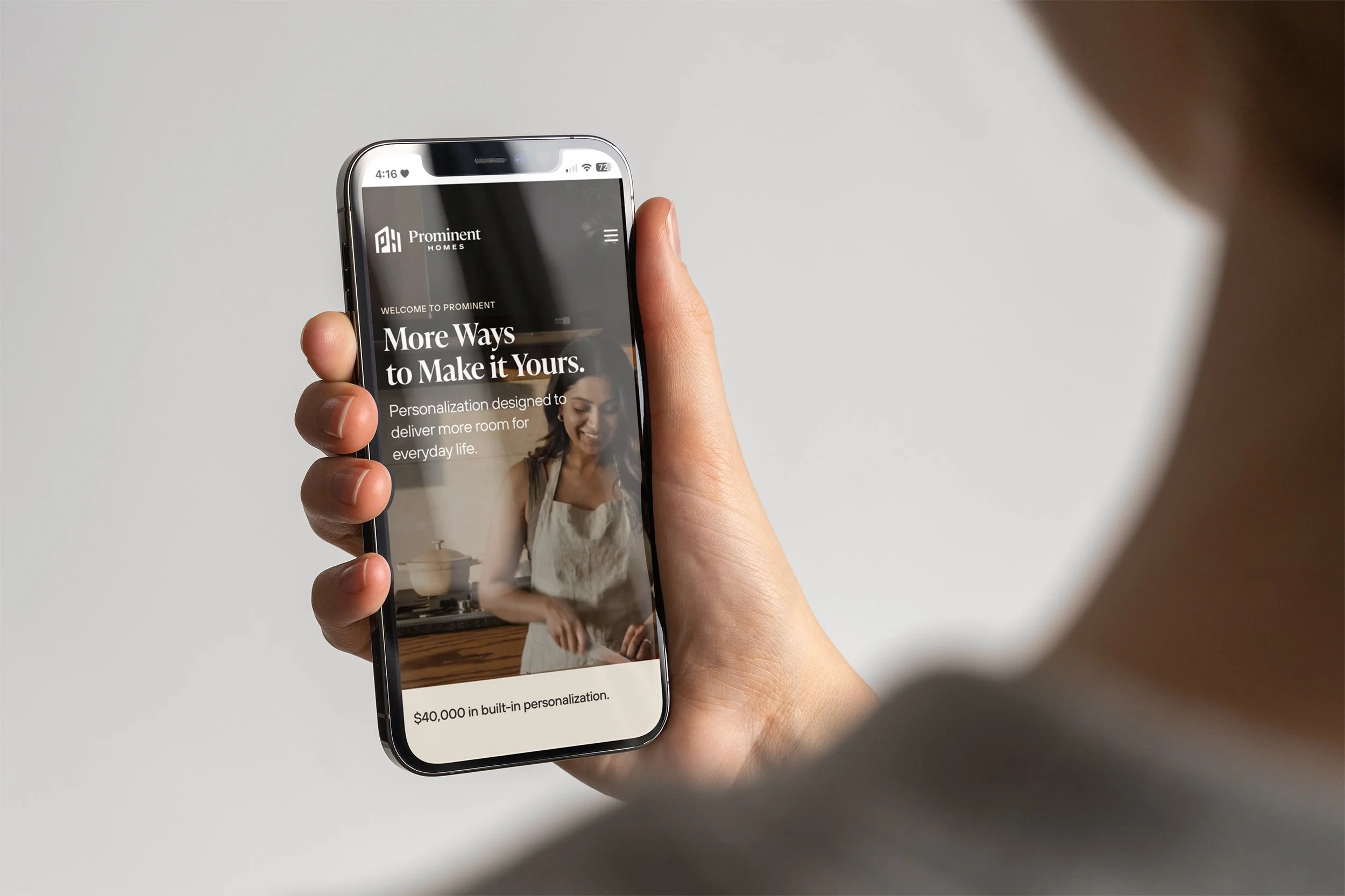 A person holding a smartphone displaying a website for Prominent Homes, showcasing personalized home features with an image of a woman in a kitchen.