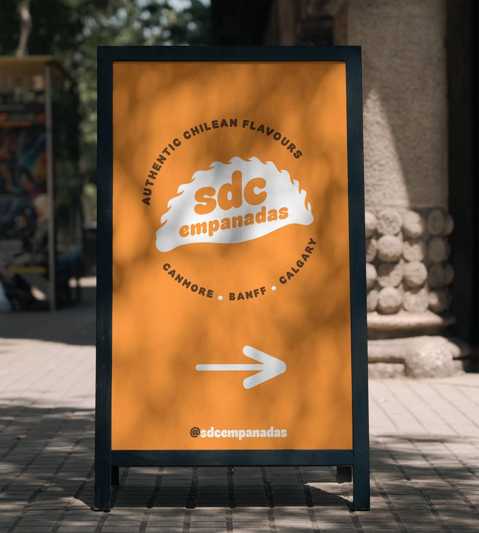 Signboard advertising empanadas with a list of Argentine regions. The text reads "Authentic Chilean Flavours," "sd empanadas," and includes the Instagram handle @sdempanadas. The signboard is outdoors on a paved area with a blurred background of trees and building structures.