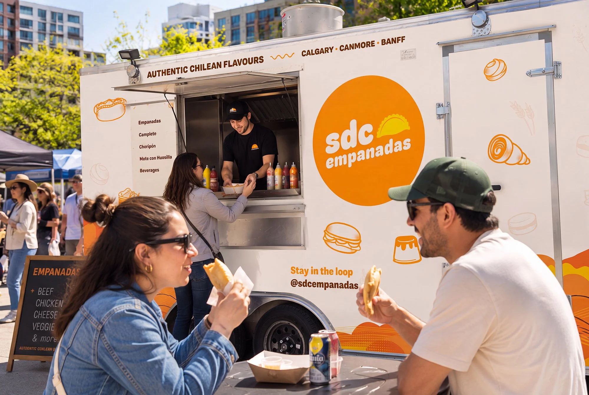 People enjoying empanadas in front of a food truck called SD Empanadas at an outdoor event, with the truck offering authentic Chilean flavors and various menu options. The scene includes a diverse group of customers eating and interacting, with the food truck decorated with orange and white graphics, and surrounded by trees and other tents.