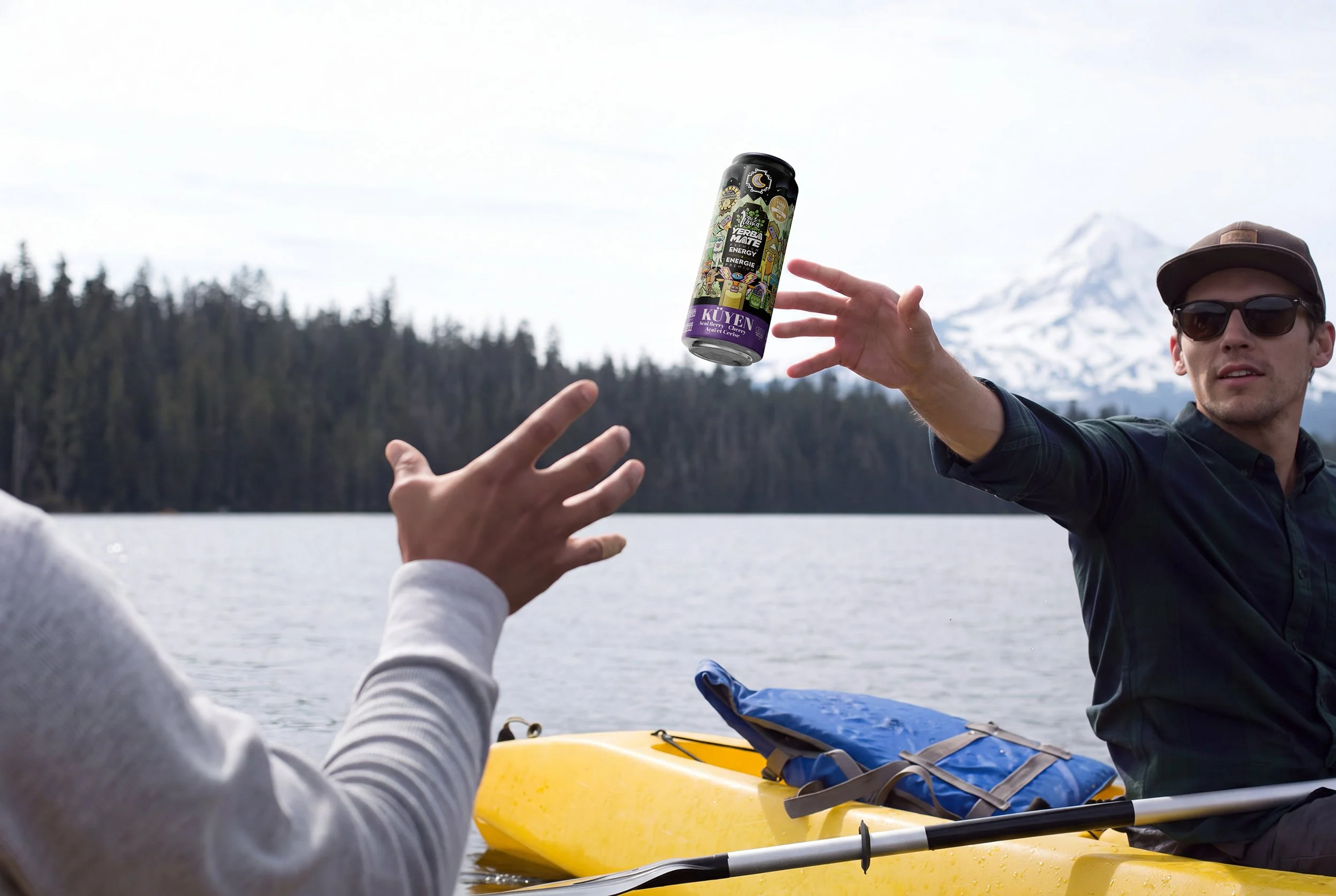 A person with sunglasses and a baseball cap reaches out to catch a can of energy drink being tossed by another person in a yellow kayak on a lake, with mountains and trees in the background.