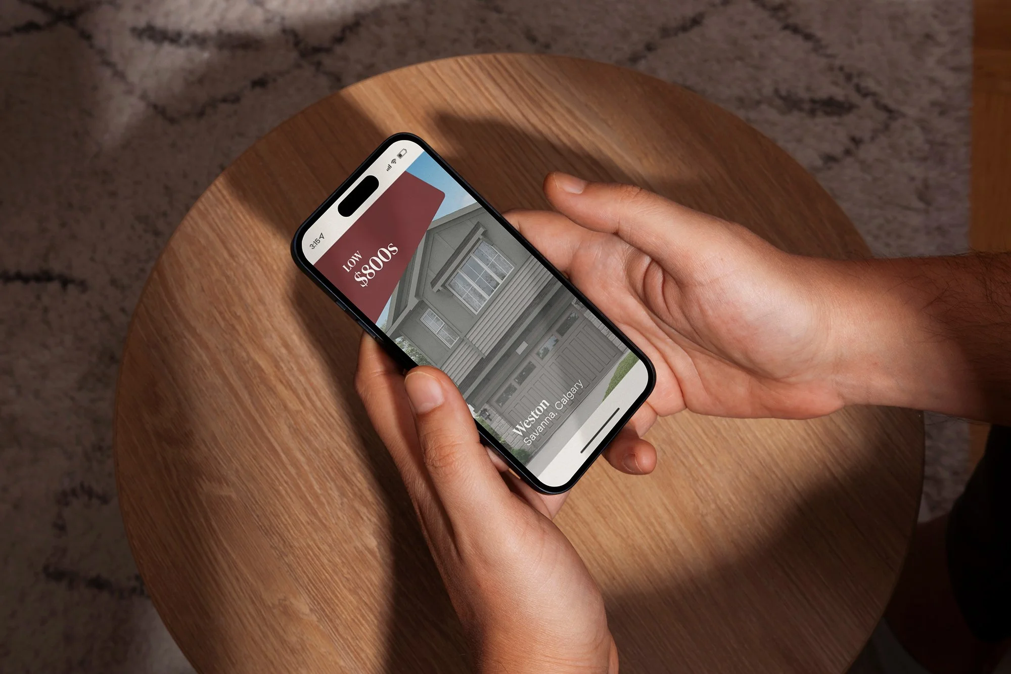 Person holding a smartphone displaying a real estate app with house images and prices, sitting at a wooden table.