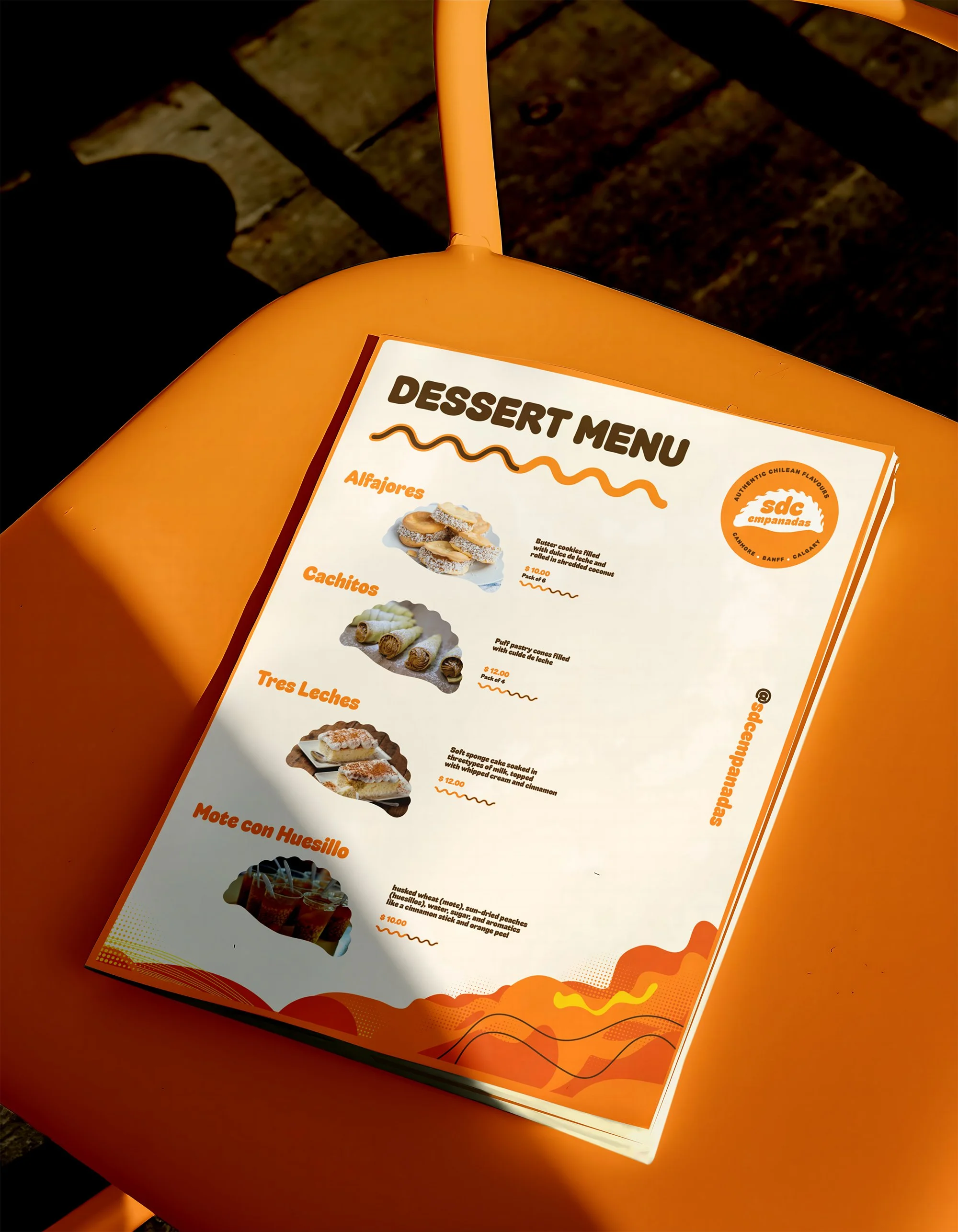 Close-up of a dessert menu on an orange table, featuring images and descriptions of Colombian-style desserts like alfajores, cachitos, tres leches, and mote con huesillo.