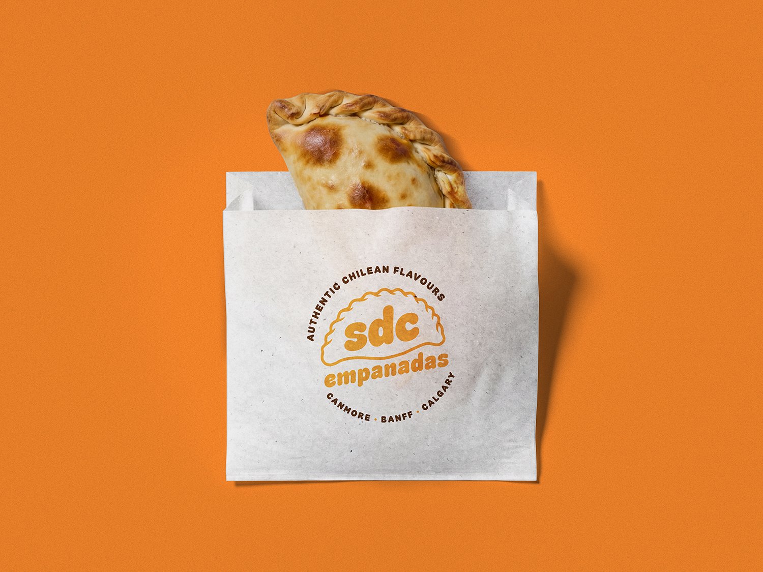 A white paper bag with a logo reading 'sdc empanadas' and the text 'Authentic Chilean Flavours, Canmore, Banff, Calgary' printed on it. An empanada with a golden-brown crust and crimped edge is sticking out of the bag, set against an orange background.