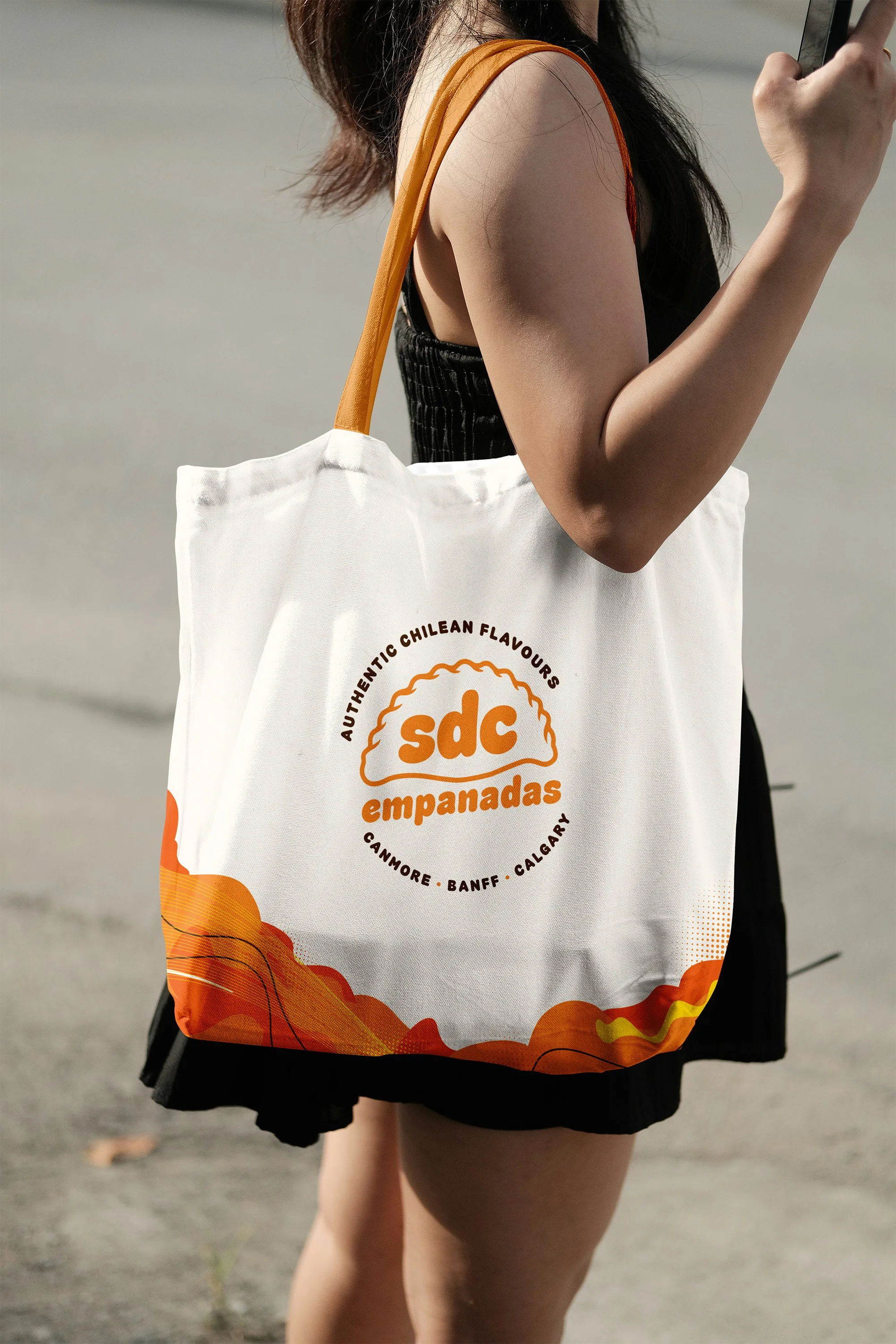 A woman wearing a black dress with orange straps, carrying a white tote bag with orange text and design, standing outdoors on a gray pavement.