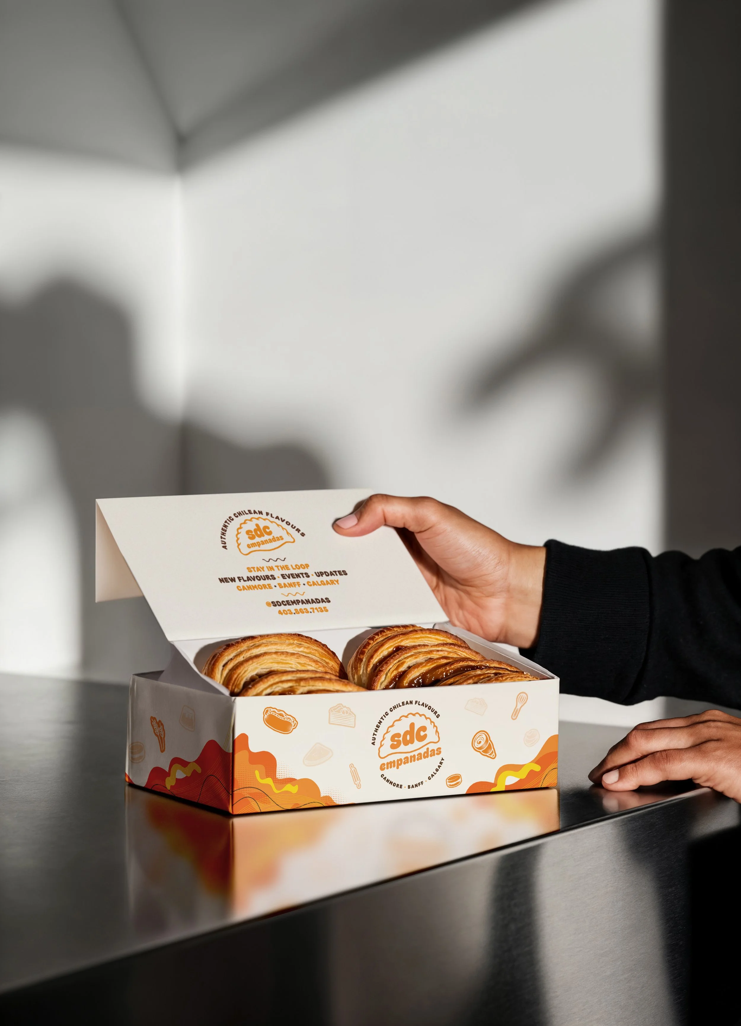 A person opening a box of empanadas with the logo 'SDC Empanadas' on the inside lid, featuring a hand with dark skin and black sleeve. The empanadas are golden brown and placed inside the box on a reflective metallic surface, with a blurred gray background and shadows.