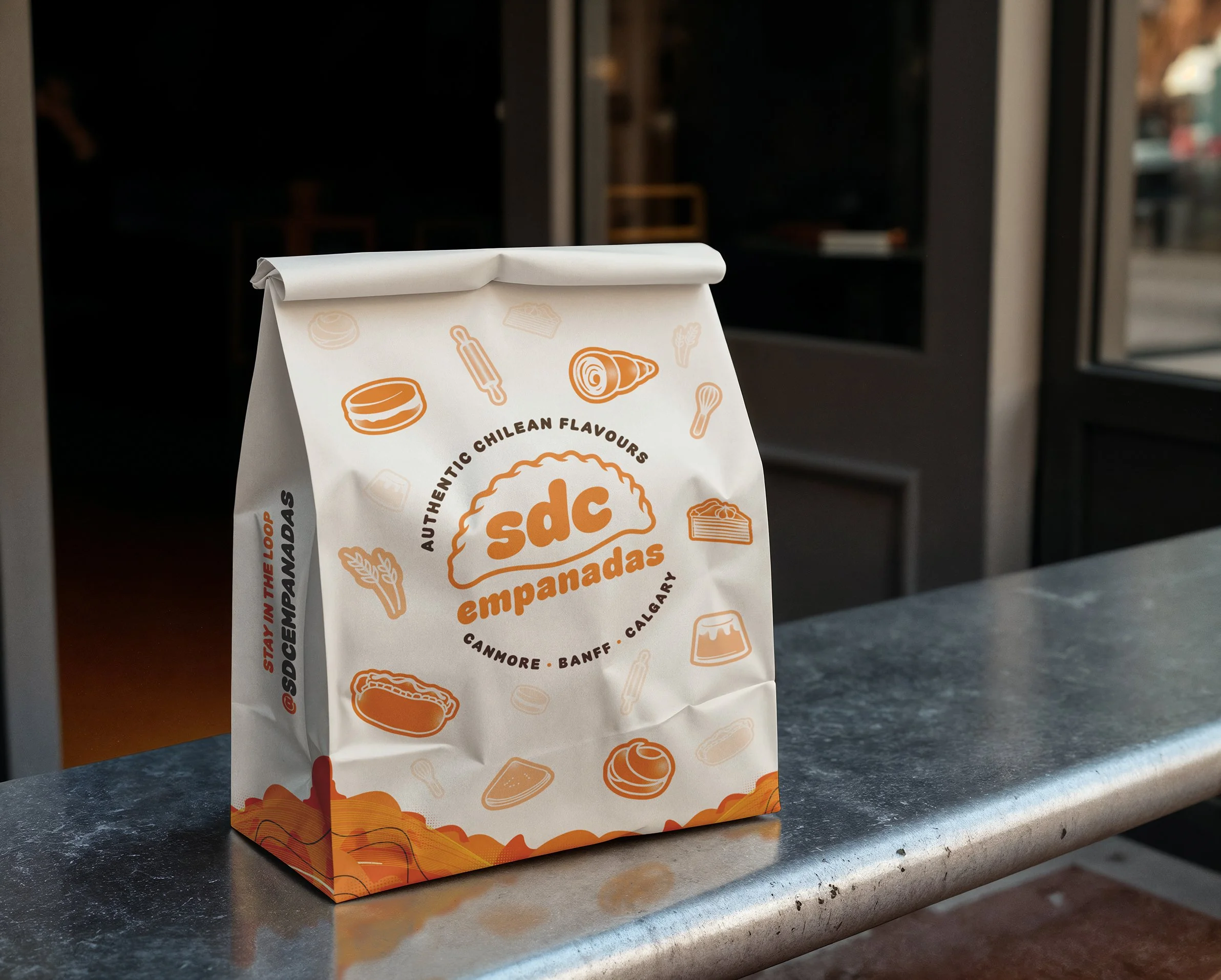 A white paper bag with orange illustrations and black text, sitting on a dark countertop near a window, with a blurred background.