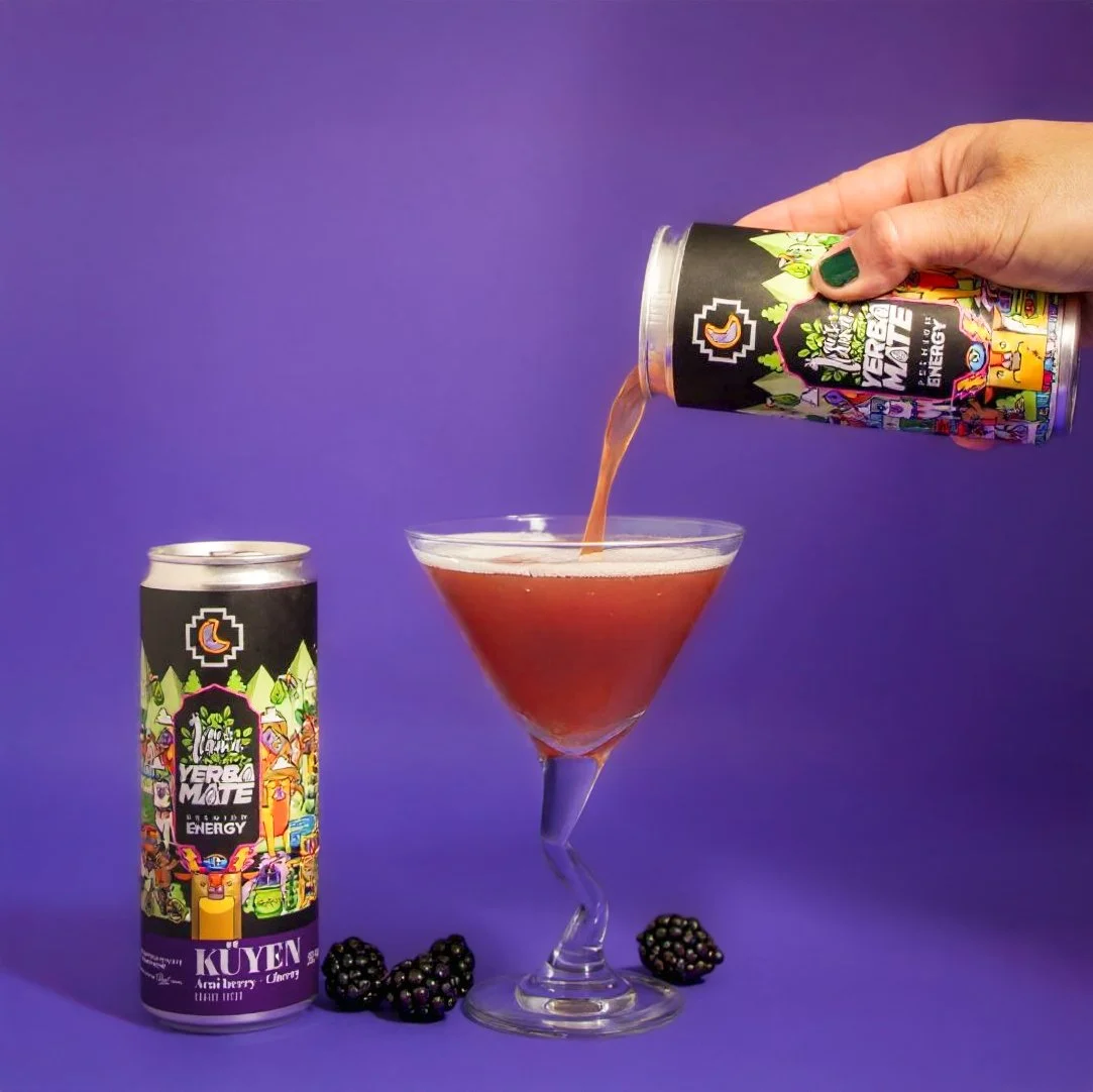A hand pours a berry-flavored energy drink from a colorful can into a cocktail glass, with a can and blackberries on a purple background.
