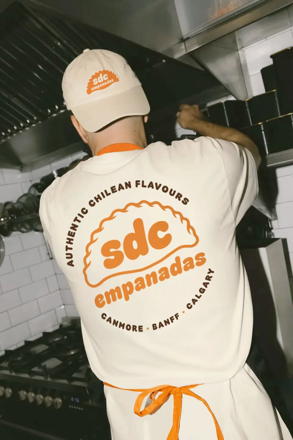 A chef wearing a beige hat and white apron with orange text prepares food in a commercial kitchen.