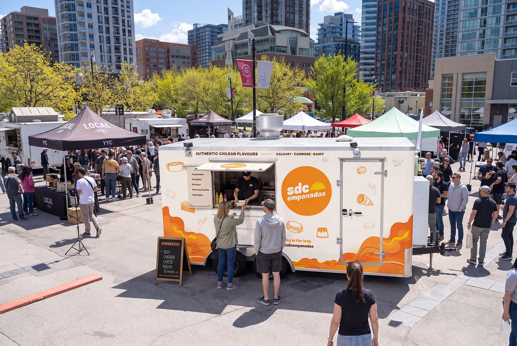 Outdoor market with food trucks and tents set up in an urban plaza surrounded by tall buildings. A food truck labeled "sdC empanadas" serves Chilean-style empanadas, and there are many people shopping and socializing.