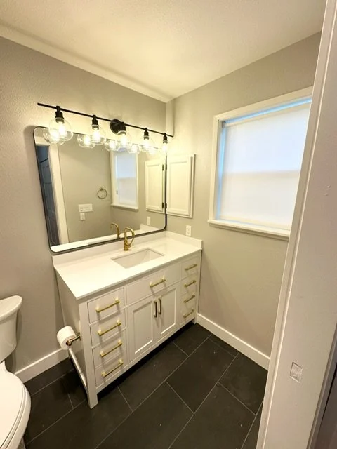 Bathroom vanity with white cabinet, gold hardware, and a white countertop with a built-in sink. Above the vanity is a large mirror with three light bulbs. To the right is a window with a white blind, and there is a toilet in the corner.
