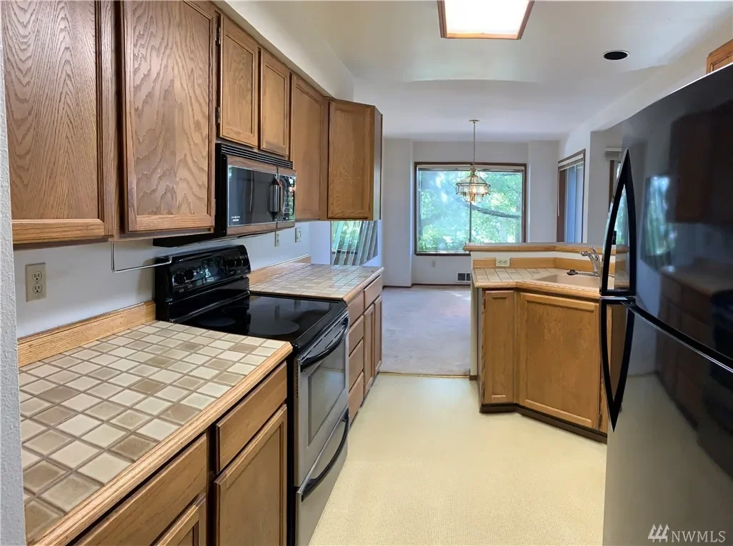 Kitchen with wooden cabinets, black stove, microwave, refrigerator, tiled countertops, and a view into a dining area with a window and hanging light fixture.