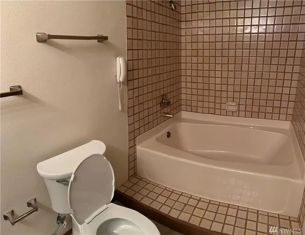 Bathroom featuring a toilet with the lid open, a wall-mounted telephone, and a bathtub surrounded by brown and beige small square tiles.