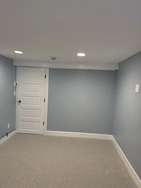 Empty interior room with light blue walls, beige carpet, a closed white door, and recessed ceiling lights.