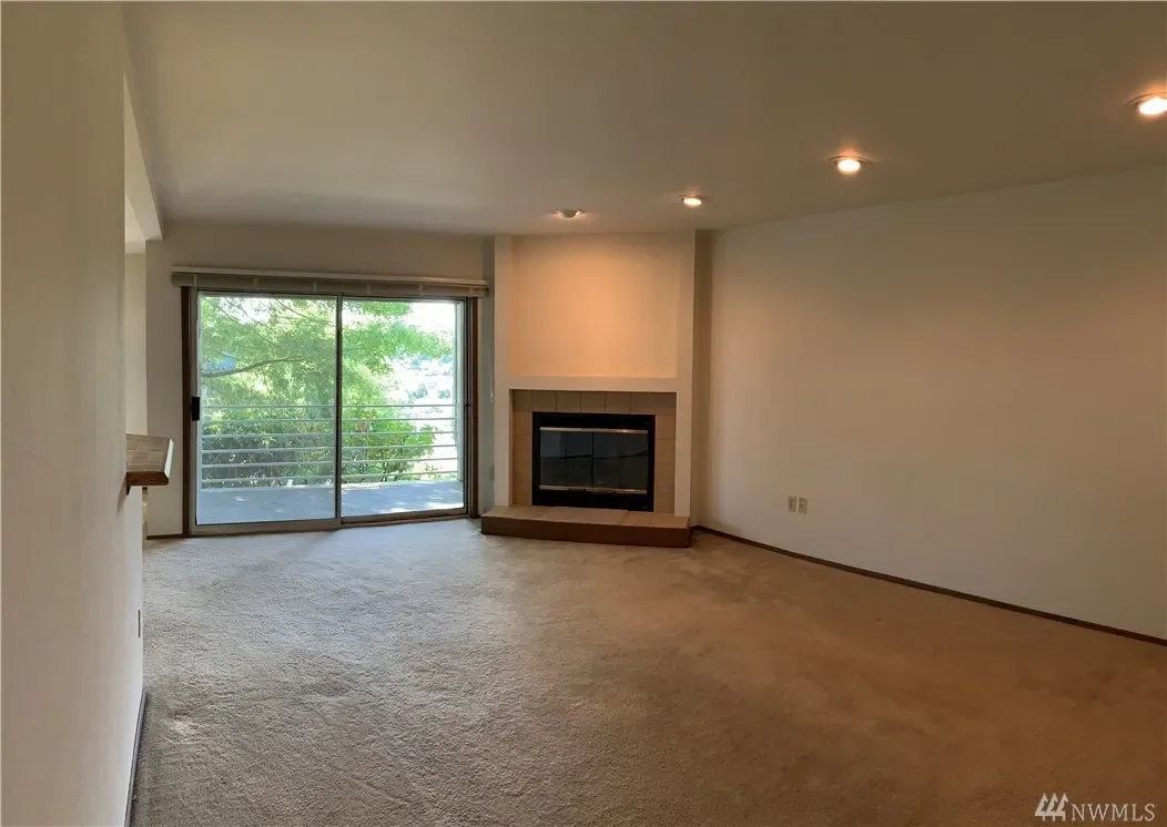 Empty living room with beige carpet, white walls, a sliding glass door leading to a balcony, and a fireplace with a simple beige mantel.