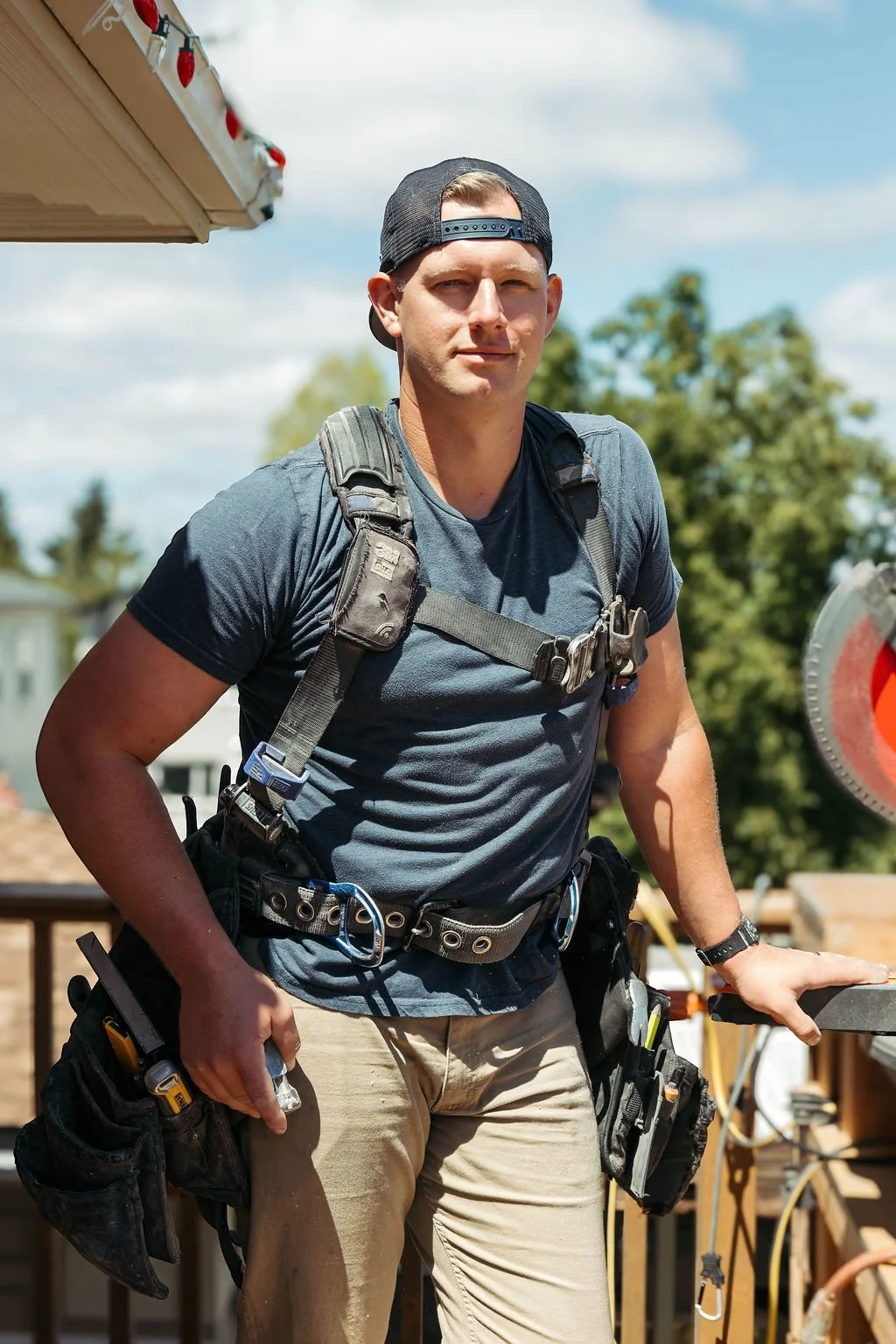A young man wearing a backwards black baseball cap, gray t-shirt, and beige work pants stands outdoors with a serious expression. He has a tool belt, a backpack harness, and various tools attached to his waist and pockets. The background shows a partly cloudy sky and green trees.