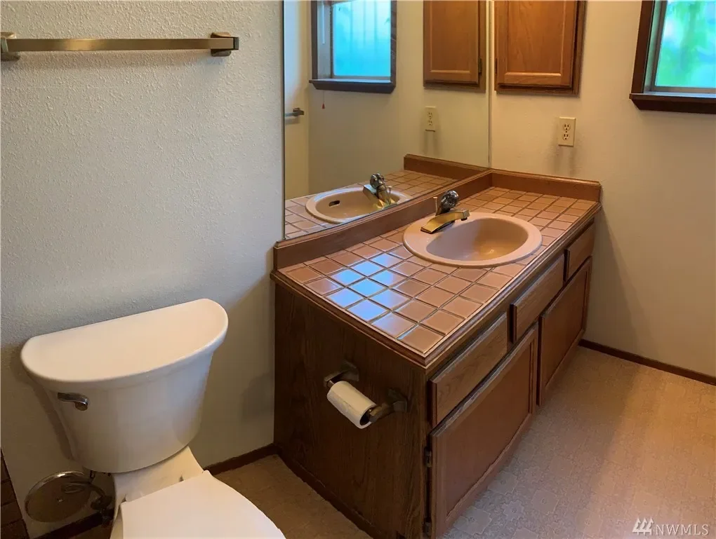 A small bathroom with a toilet, a wooden vanity with a tile countertop, a sink, and a large mirror. There is a window with a view outside and a towel bar on the wall.