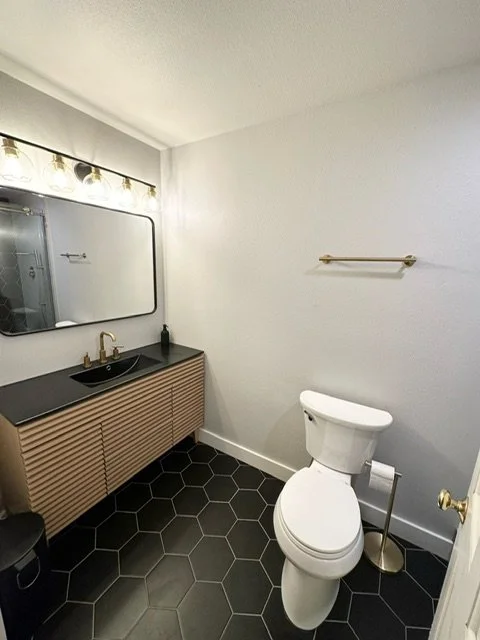 Bathroom with white walls, a floating vanity with a black countertop and sink, a large rectangular mirror, a toilet, a towel rack, and black hexagonal floor tiles