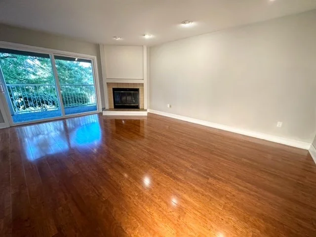 Empty living room with wooden floors, a fireplace, and sliding glass doors leading to a balcony overlooking trees.