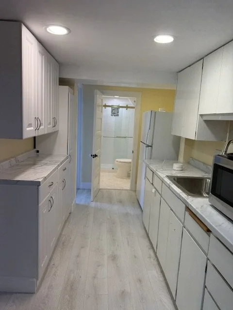 Kitchen with white cabinets, marble countertops, a refrigerator, microwave, sink, and view into a bathroom with a toilet.