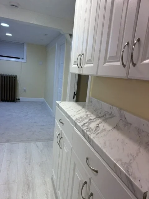 Interior view of a kitchen with white cabinets, marble countertop, beige walls, and light-colored flooring, leading to a living area with a radiator and window.