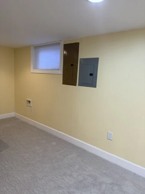 Empty room with beige walls, a small window with blinds, electrical boxes, and a power outlet.