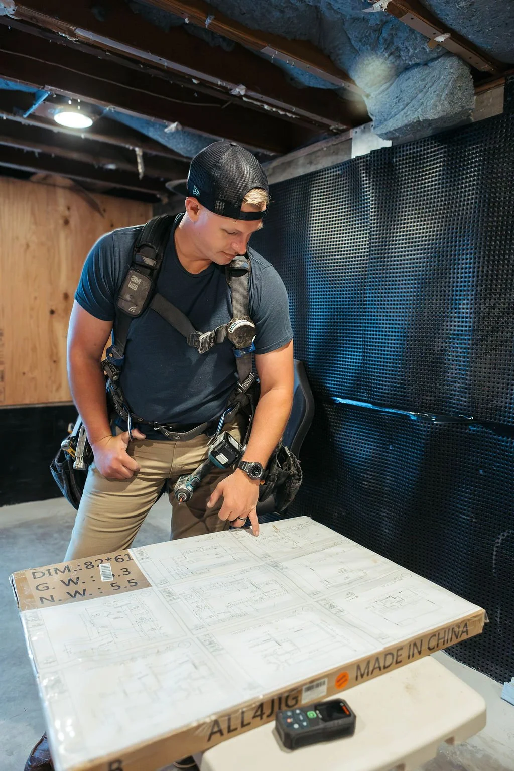 A man in work attire, wearing a black cap and backpack, is looking at and pointing to technical blueprints or architectural plans on a table in a partially finished basement or construction site.
