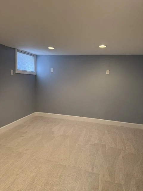 Empty room with gray walls, beige carpet, two ceiling lights, and a small window.