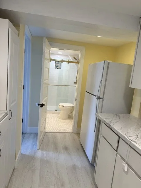 View of a small kitchen leading to a bathroom, with a refrigerator, white cabinets, and a marble countertop.