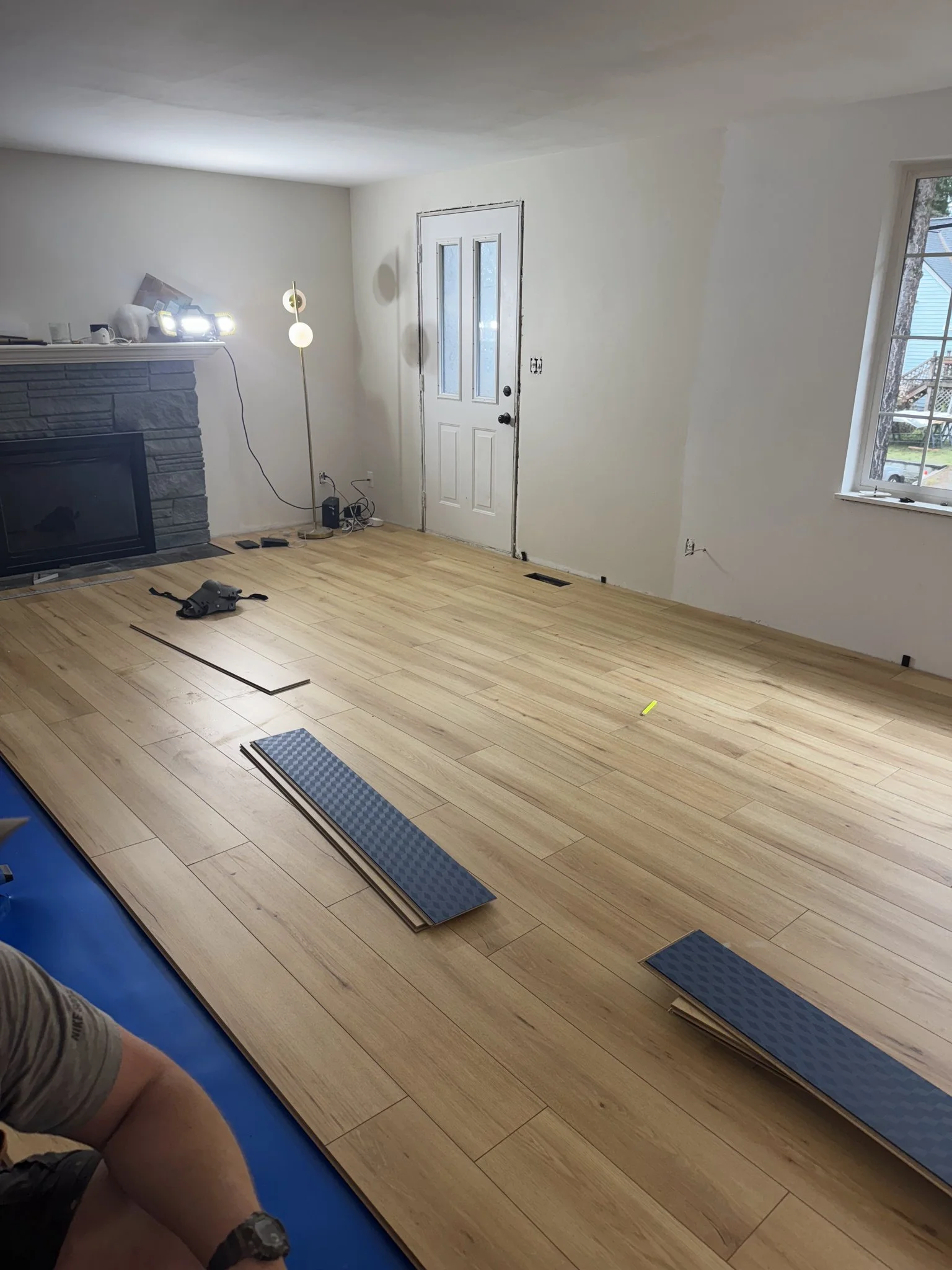 Living room with hardwood flooring, a stone fireplace, a white door, a window, and construction tools and materials scattered on the floor, indicating ongoing renovation.