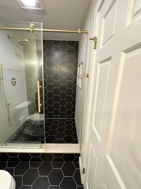 Bathroom with a walk-in shower featuring black hexagonal tiles and a glass door, and a white door on the right side.