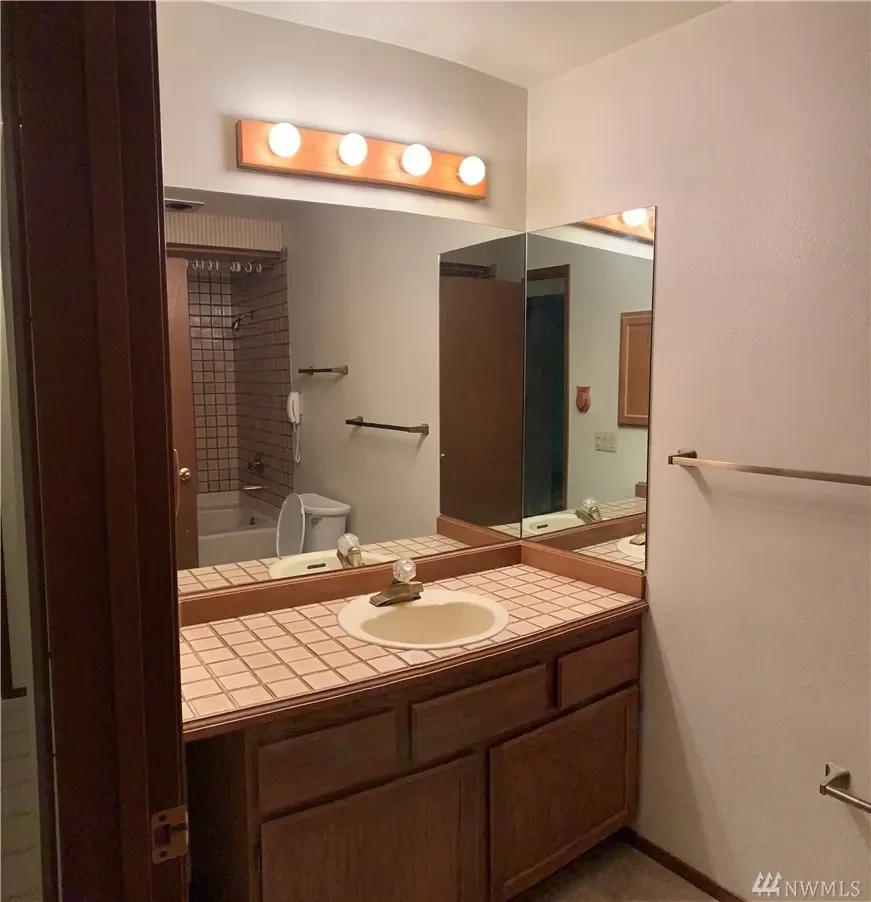 Bathroom vanity with tiled countertop, a sink, and a large mirror, with a three-bulb light fixture above, and a room visible in the background through the reflection.