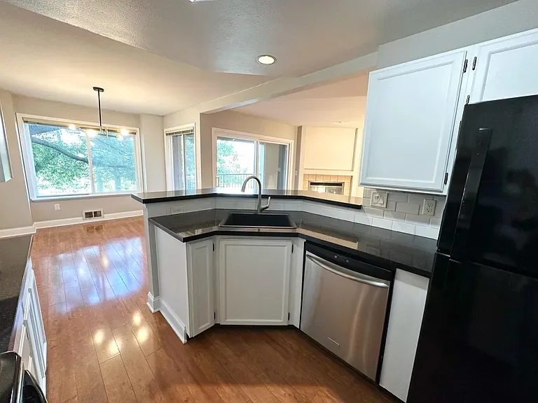 Modern kitchen with black countertops, white cabinets, stainless steel dishwasher, and black refrigerator. Open living area with large windows and hardwood floors.