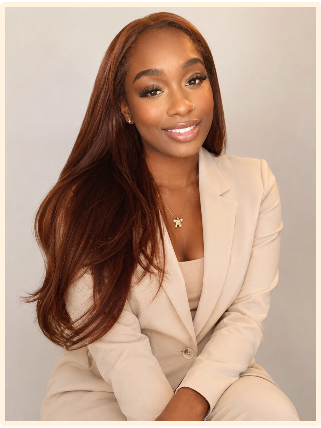 A woman with long, wavy auburn hair, wearing a beige blazer and matching top, smiling and seated against a plain light gray background.