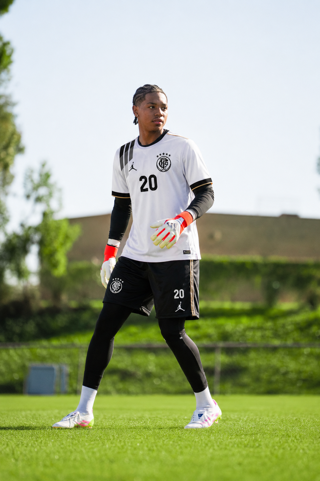 A male soccer player wearing a white and black jersey with the number 20, black shorts, and long black leggings, standing on a green soccer field with trees and a building in the background.