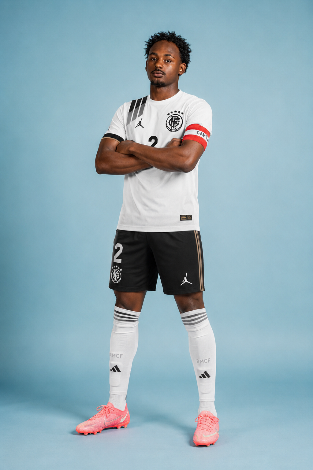 A man in a soccer uniform with arms crossed, standing against a blue background. He wears a white and black jersey with a team logo, black shorts, white socks, and pink soccer cleats.