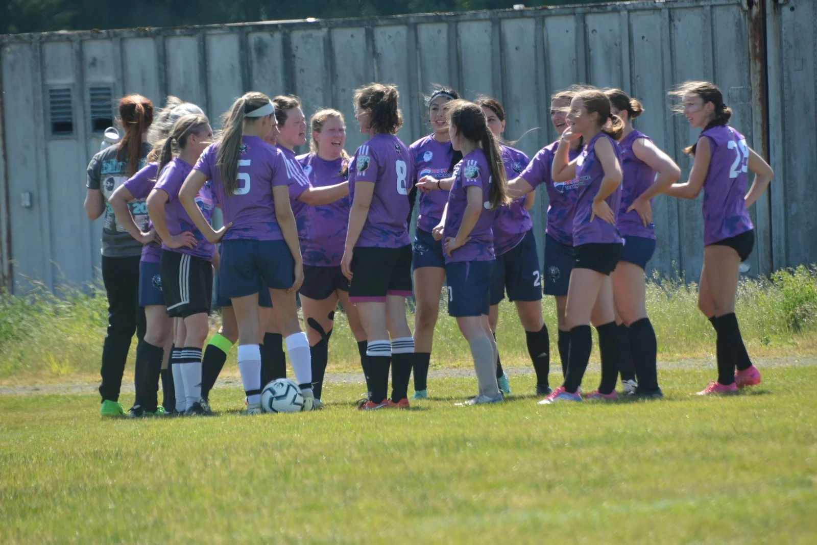 A group of female soccer players in purple jerseys standing in huddle on a grassy field, with a soccer ball at their feet, surrounded by a weathered metal fence.