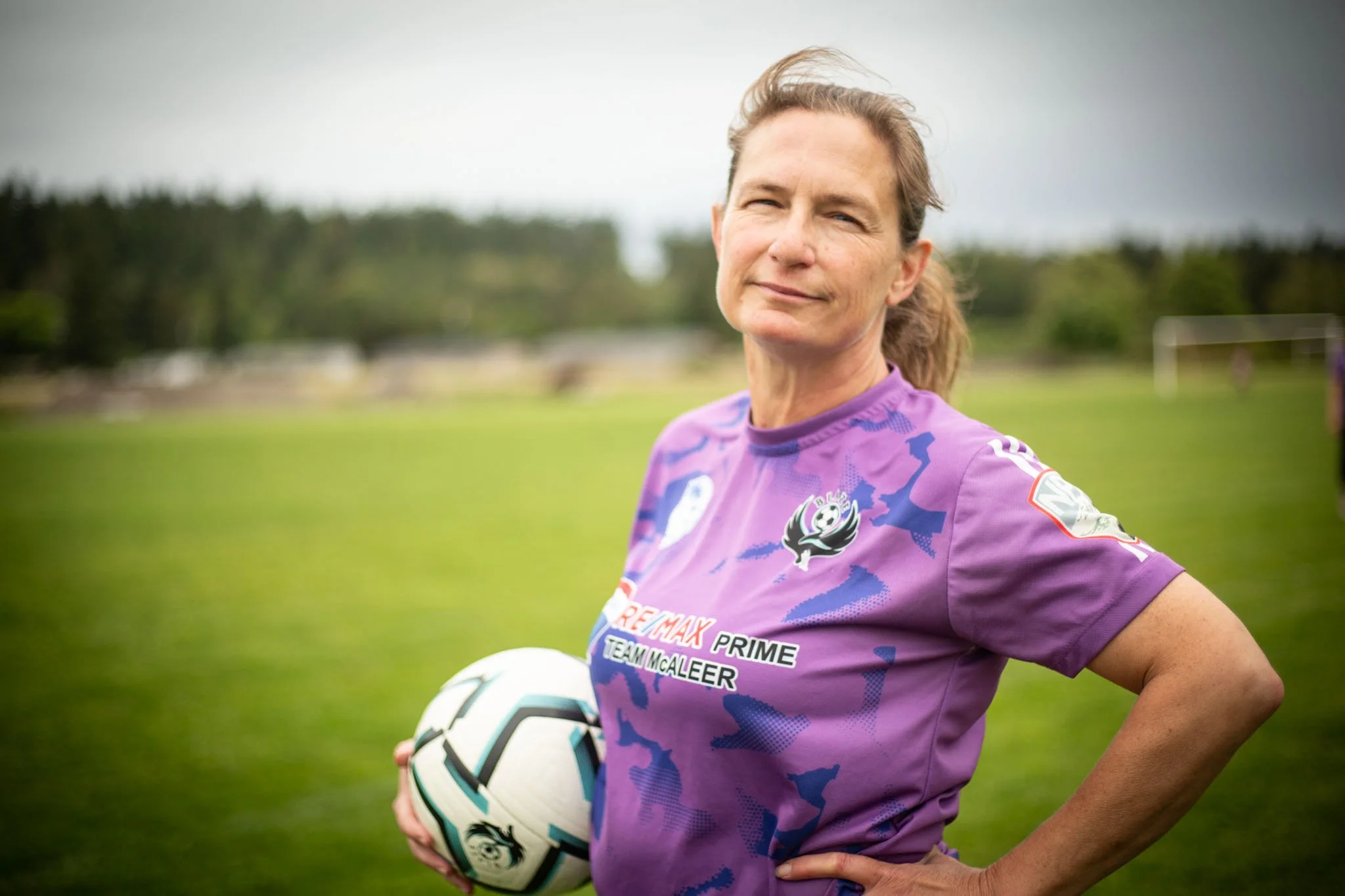 A woman in a purple soccer jersey holding a soccer ball on a grassy field.