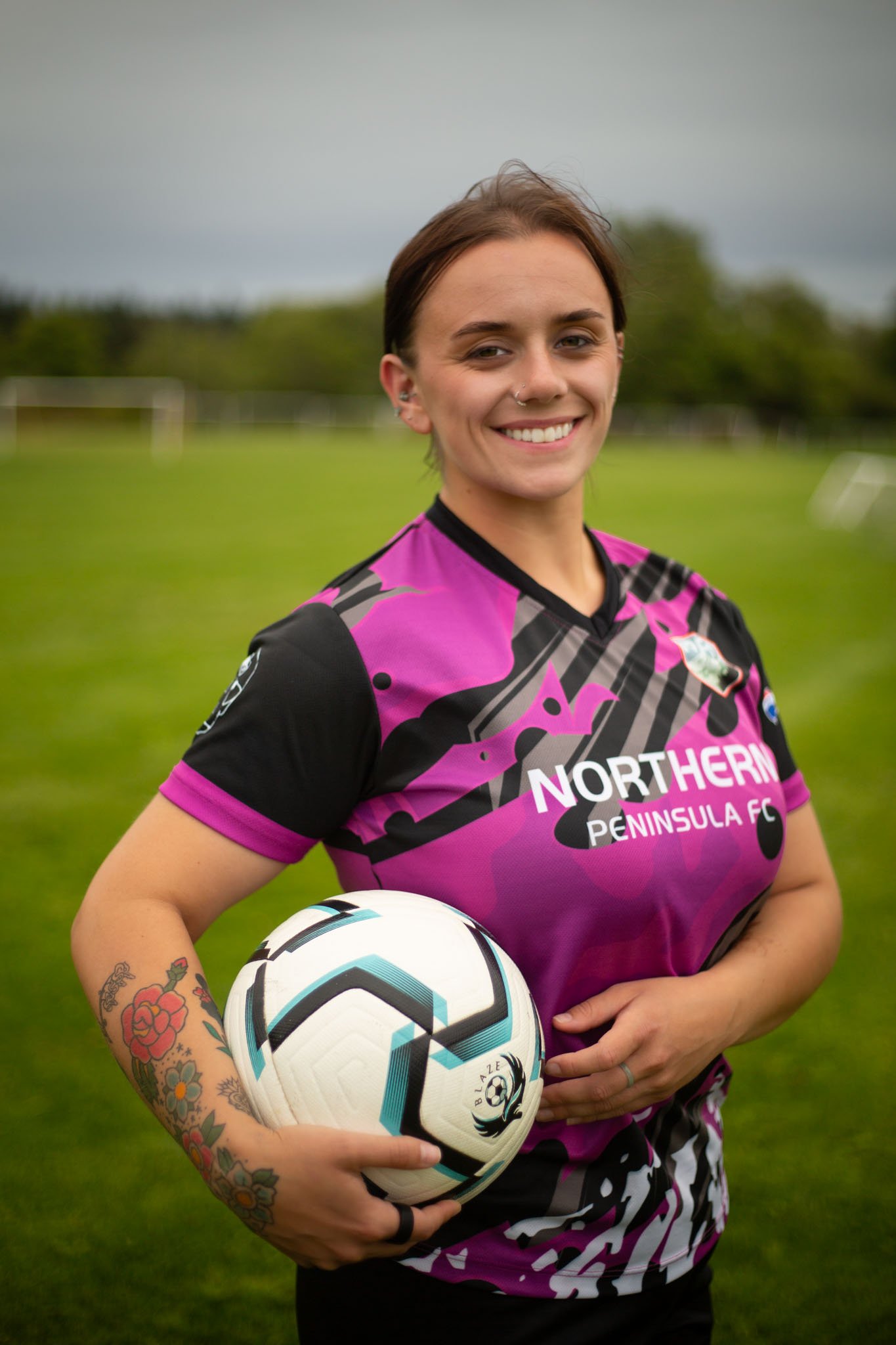 A smiling woman with tattoos on her left arm holding a soccer ball on a grassy field on a cloudy day. She is wearing a pink and black sports jersey with the text 'Northern Peninsula FC'.