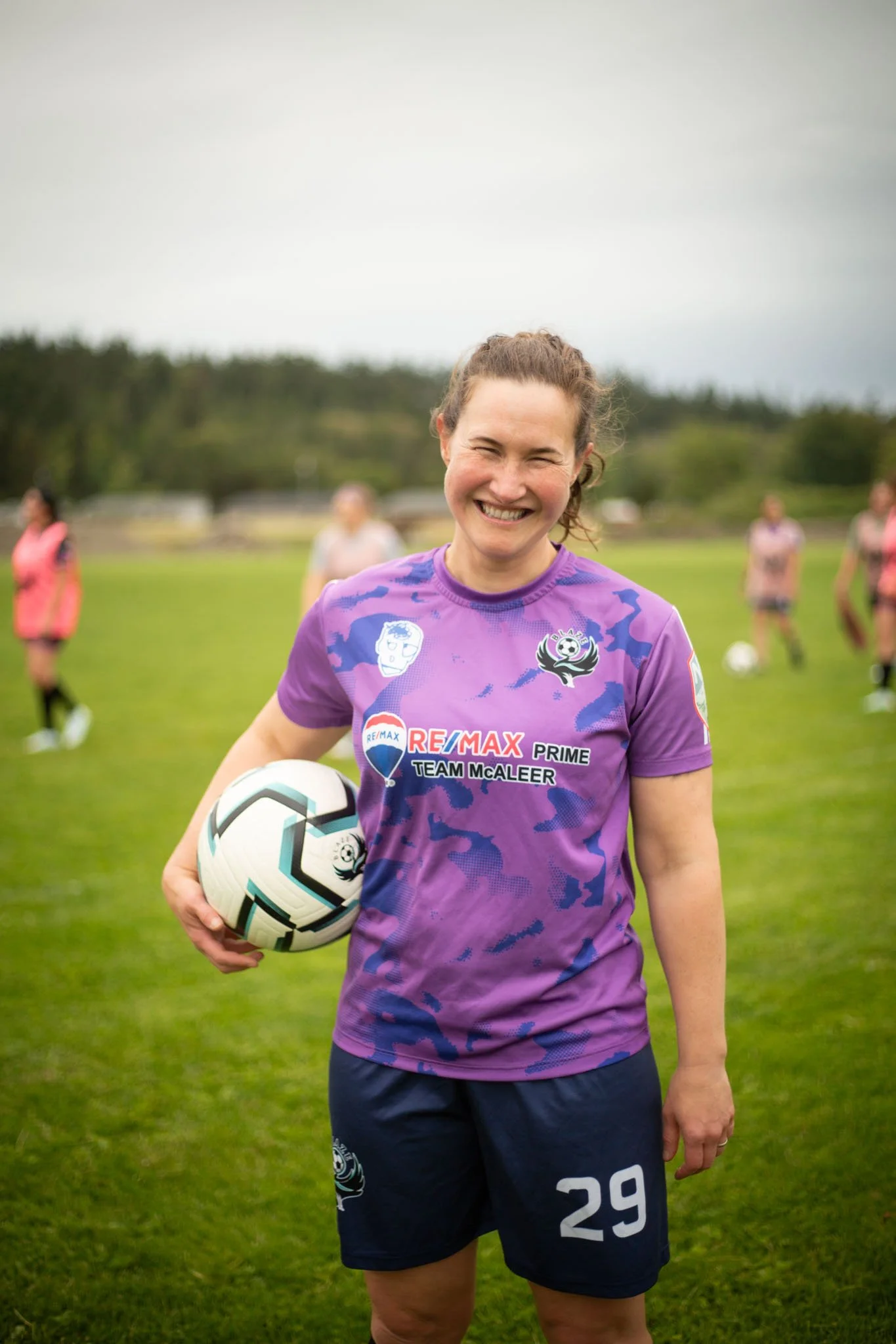 A smiling female soccer player in a purple and black uniform holding a soccer ball, standing on a soccer field with other players in the background.