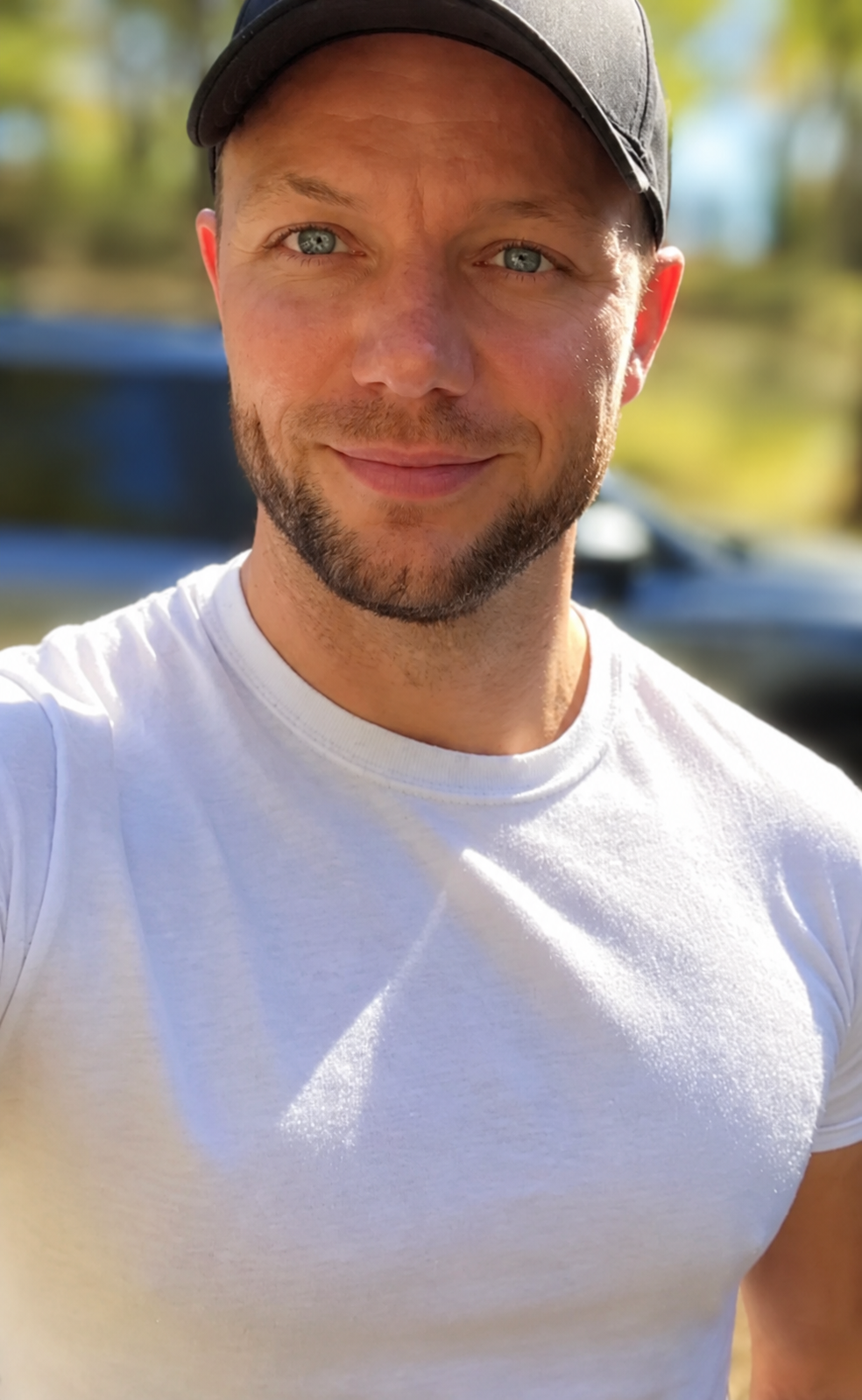 Close-up of a man with blue eyes, a beard, wearing a black cap and white t-shirt, outdoors with blurred trees and cars in the background.