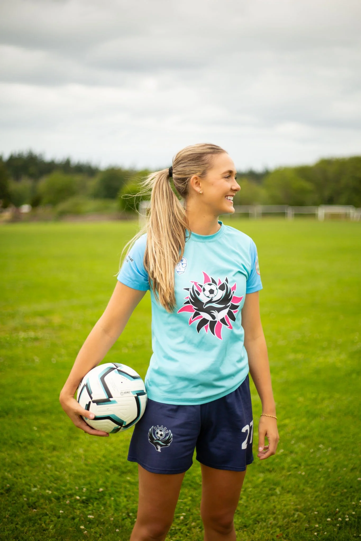 A young woman with long blonde hair in a ponytail is smiling and holding a soccer ball on a green field under cloudy skies. She is wearing a light blue sports T-shirt and navy shorts, with a logo on the shirt and shorts.