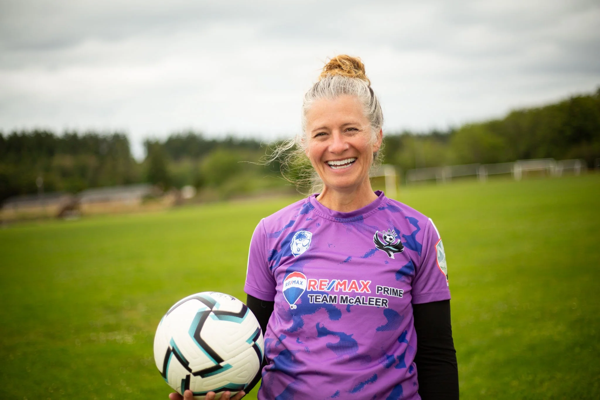 A smiling woman in a purple soccer jersey holding a soccer ball outdoors on a cloudy day.