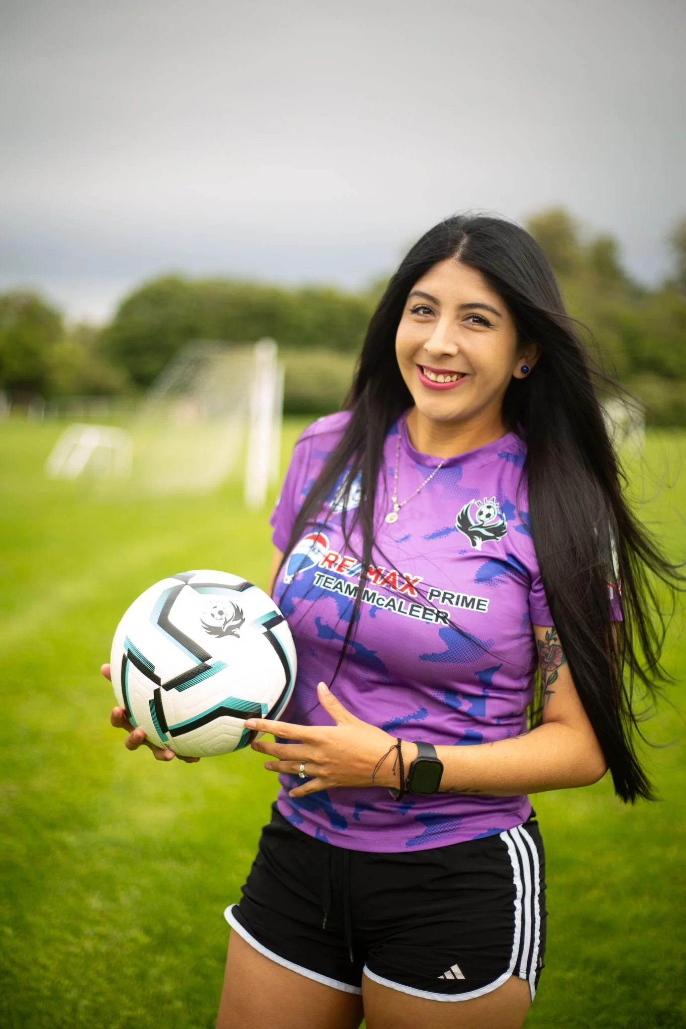 A woman in a purple sports jersey holding a soccer ball and pointing at it, standing on a grassy field with soccer goals and trees in the background.