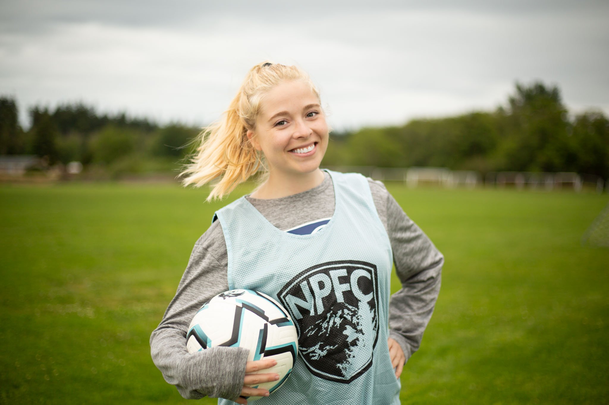 Young female soccer player smiling on the field holding a soccer ball, wearing a gray long sleeve shirt and a light blue vest, with trees and overcast sky in the background.