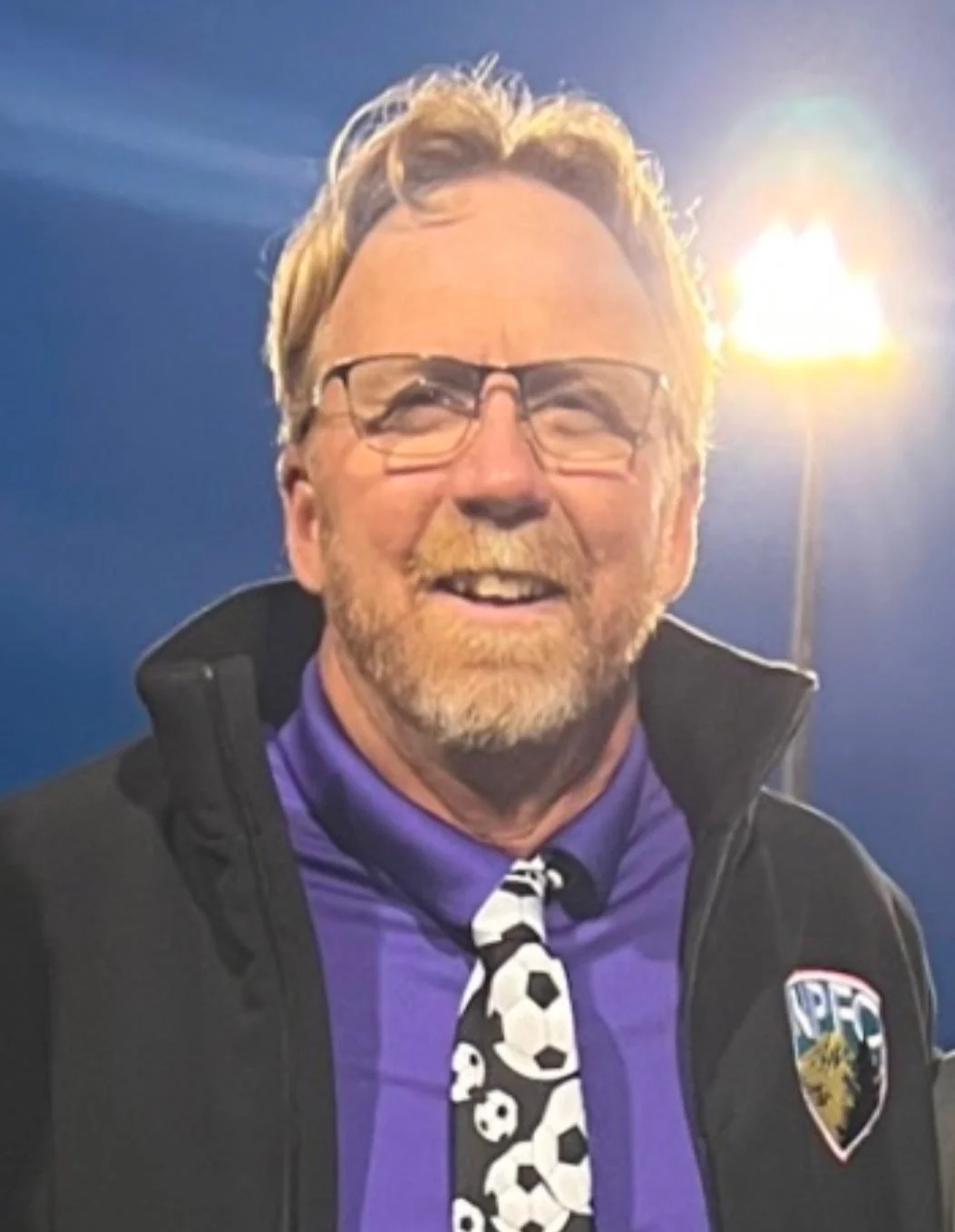 Man with glasses, a beard, and a mustache smiling, wearing a black jacket, purple shirt, and a tie with soccer ball patterns, standing outdoors with bright light behind him.