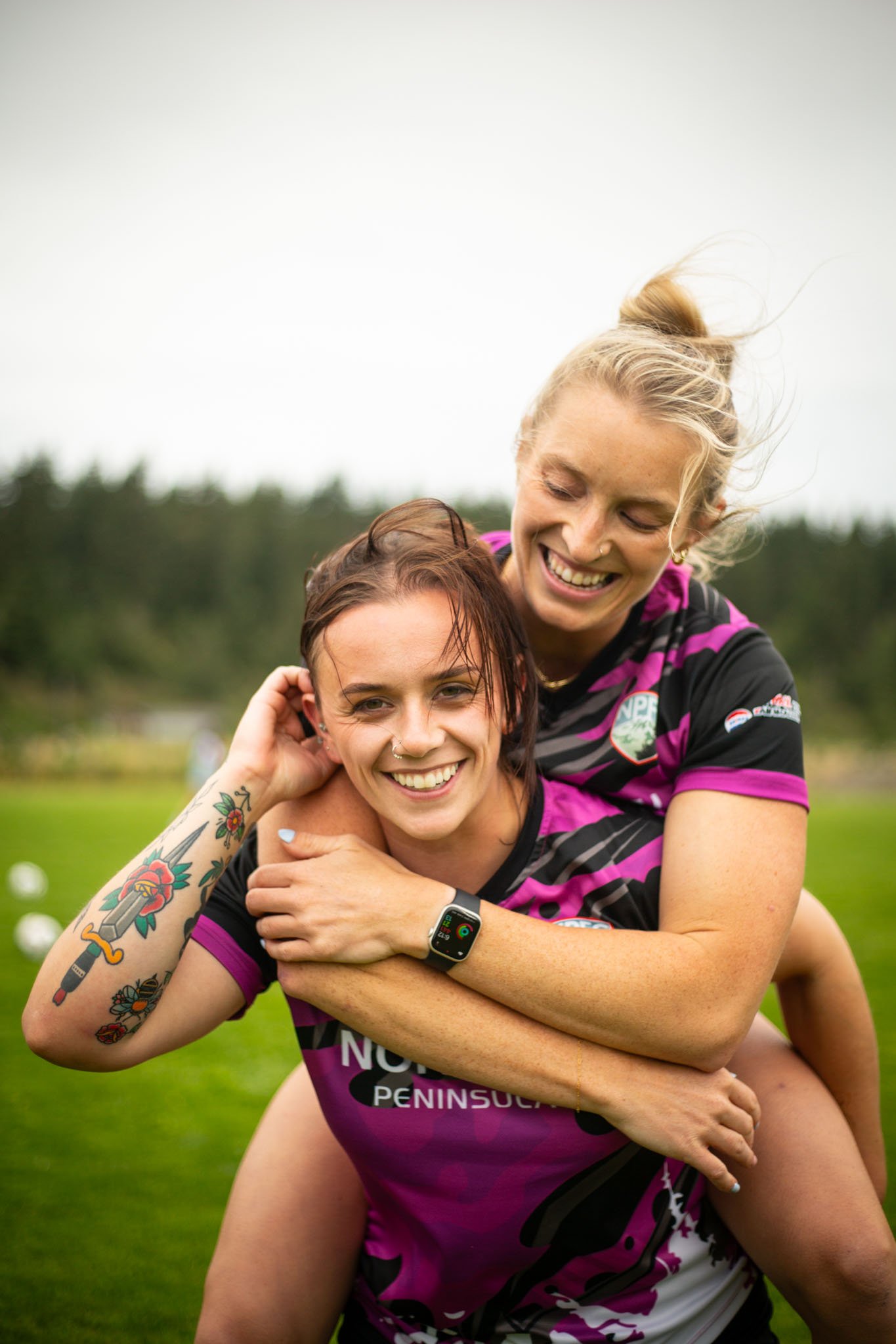 Two women celebrating outdoors on a grassy field with trees in the background, one giving the other a piggyback ride, both smiling and wearing sports jerseys.