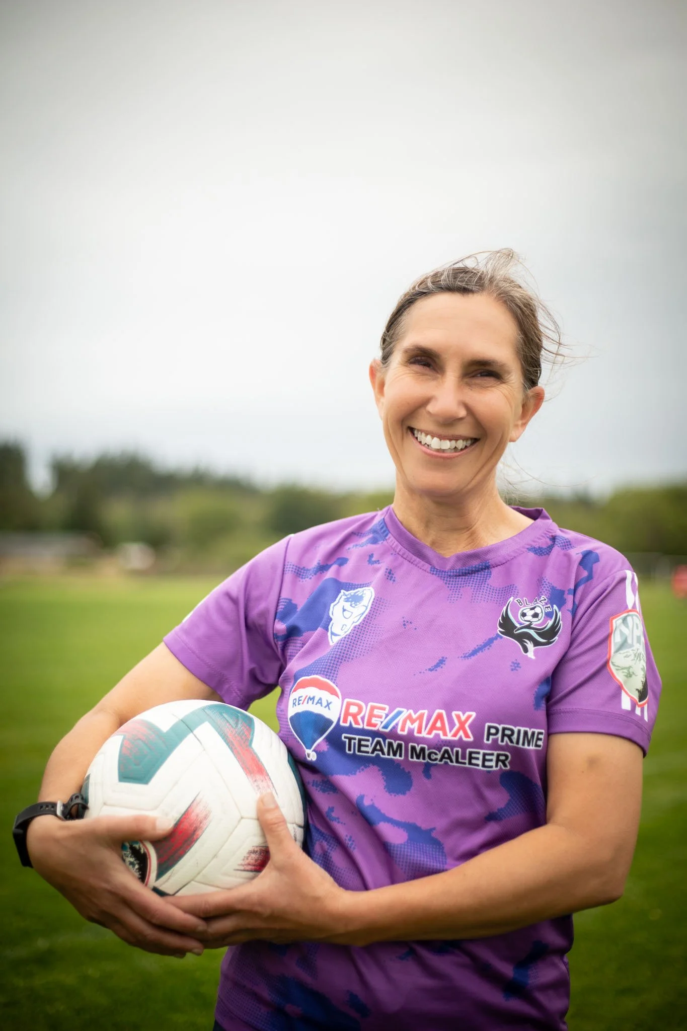 A woman wearing a purple sports jersey with sports logos, holding a soccer ball outdoors on a grassy field, smiling.