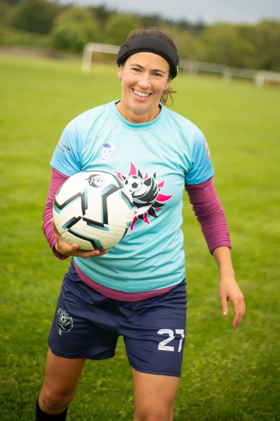 A woman on a soccer field smiling and holding a soccer ball, dressed in sports attire.