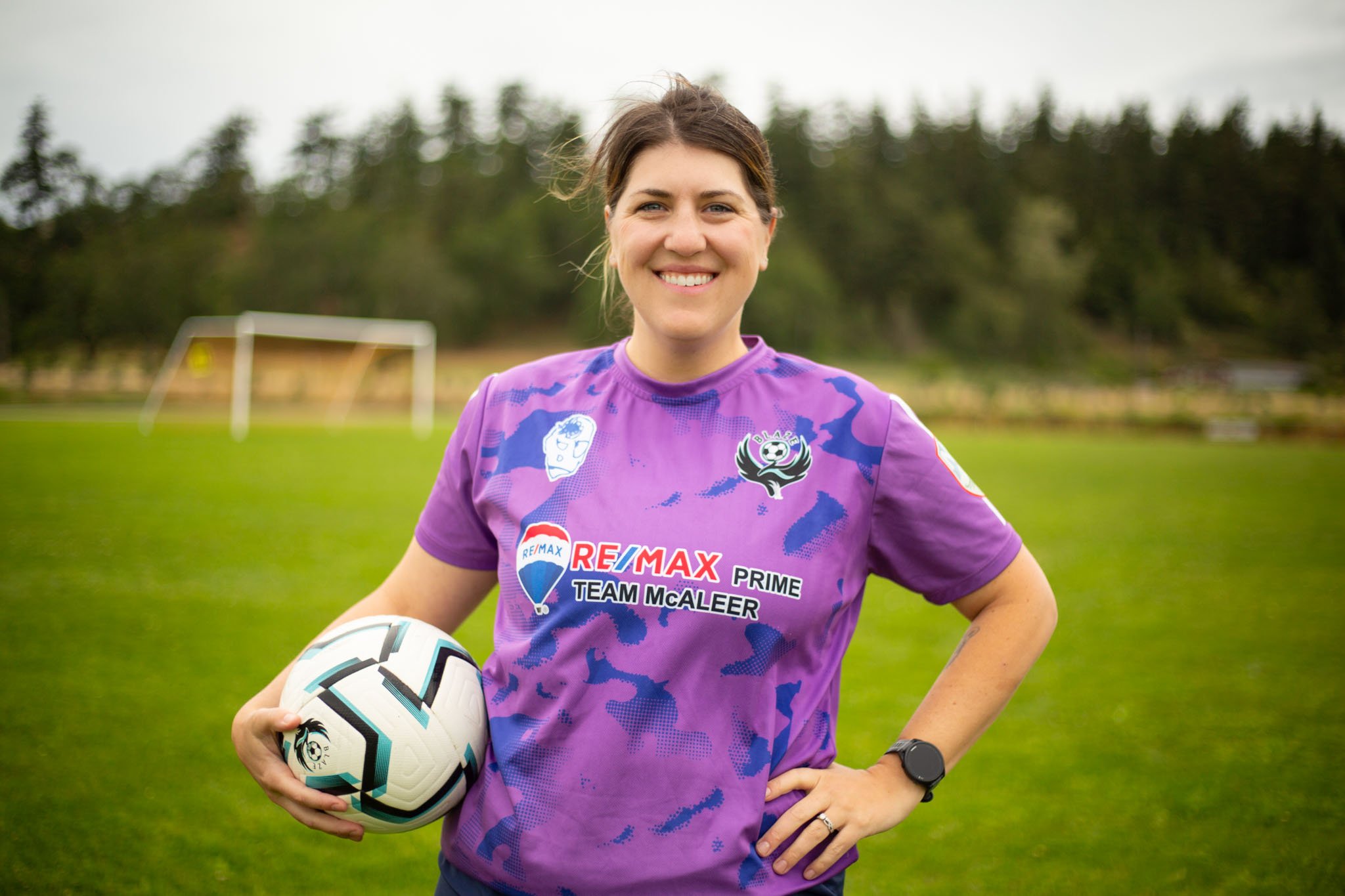A woman wearing a purple and blue sports jersey holding a soccer ball on a grassy field with trees and goalposts in the background.